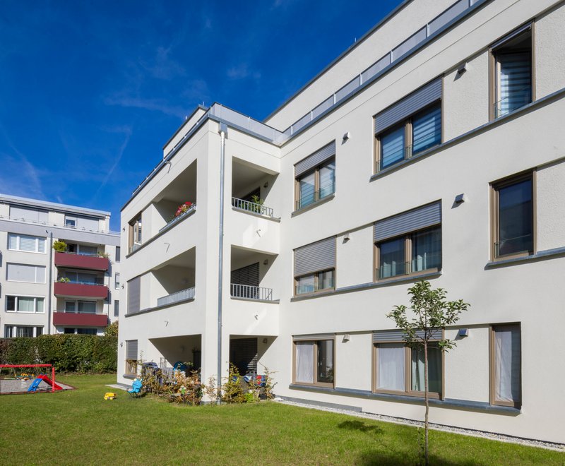 Mehrfamilienhäuser mit Balkonen Mehrere moderne, weiße Mehrfamilienhäuser mit Balkonen und großem grünen Garten im Vordergrund, bei sonnigem, blauem Himmel.