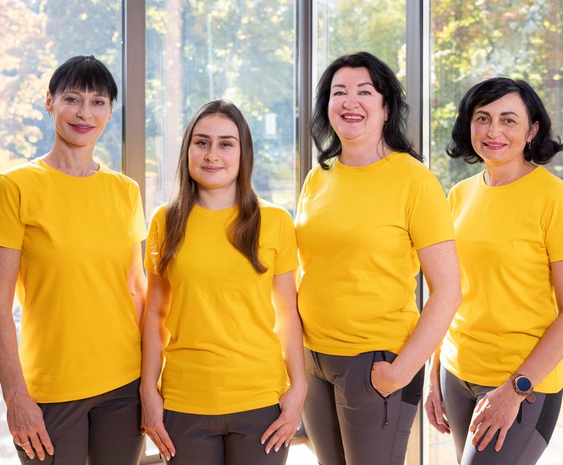 Team der SWG Vier lächelnde Frauen in gelben T-Shirts stehen nebeneinander vor großen Fenstern mit Blick auf herbstliche Bäume.