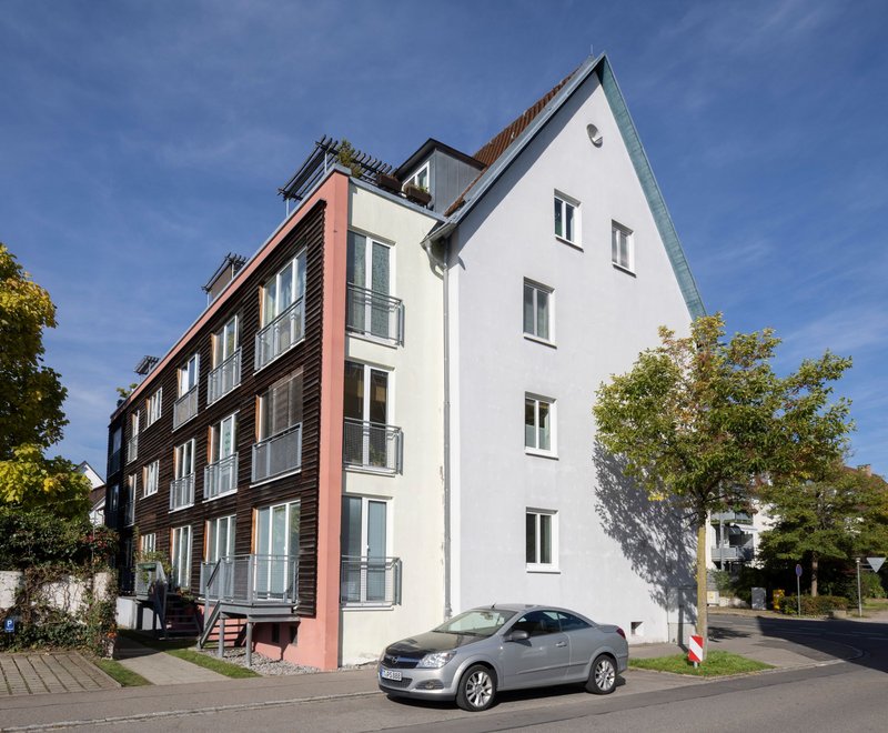 Eberhardstraße 45 Friedrichshafen Mehrstöckiges modernes Wohngebäude mit weißer Fassade und Holzverkleidung, umgeben von Bäumen. Vor dem Haus steht ein silberner Pkw. Himmel ist klar und blau.