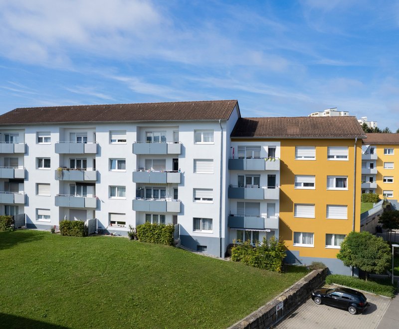 Wohngebäude mit Balkon Mehrere moderne Wohngebäude mit Balkonen, grünem Rasen und Parkplätzen an einer ruhigen Straße an einem sonnigen Tag.