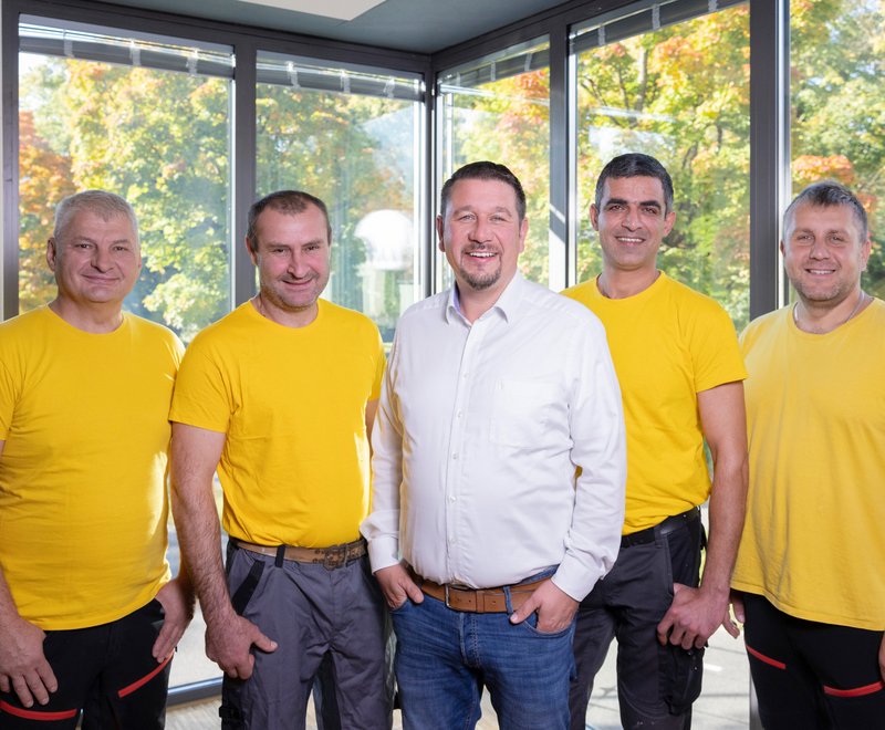 Team der SWG Friedrichshafen Fünf Männer stehen nebeneinander und lächeln. Vier tragen gelbe T-Shirts und Arbeitshosen, einer in der Mitte trägt ein weißes Hemd und Jeans. Die Szene vermittelt Teamarbeit und Freundlichkeit.