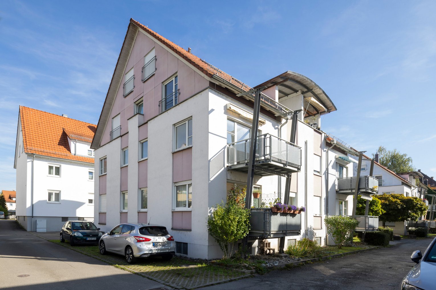 Mehrstöckiges Wohnhaus Mehrstöckiges Wohnhaus mit weißen und rosa Fassaden, mehreren Balkonen und parkenden Autos auf hell erleuchteter Straße bei blauem Himmel.