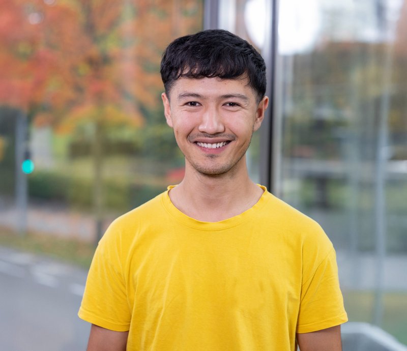 Kanmaierjiang Yimamuyushan Junger Mann mit kurzem, dunklem Haar und gelbem T-Shirt lächelt freundlich in die Kamera, herbstliche Bäume im unscharfen Hintergrund.