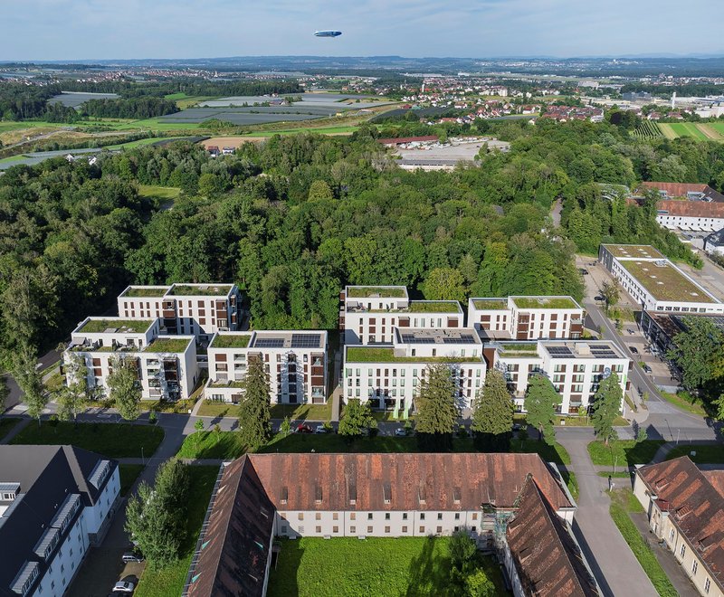 Luftaufnahme des Wohnquartiers Fallenbrunnen Luftaufnahme eines modernen Wohngebiets mit mehreren fünfstöckigen Gebäuden, umgeben von viel Grün und Bäumen. Im Hintergrund sind Felder und eine Stadtlandschaft sichtbar. Ein Zeppelin schwebt am Himmel über der Landschaft.