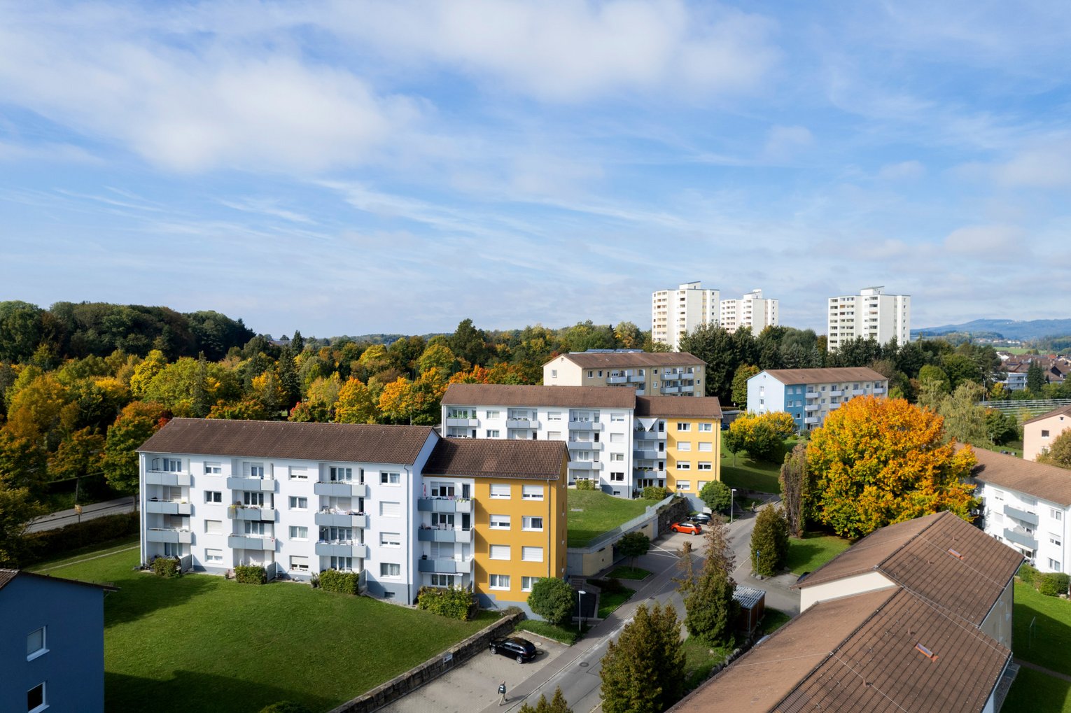 SWG Friedrichshafen Mehrere moderne Wohngebäude umgeben von Bäumen mit herbstlich gefärbtem Laub und einer grünen Wiese unter blauem Himmel mit vereinzelten Wolken. Im Hintergrund sind weitere Wohnhochhäuser und Hügel zu sehen.