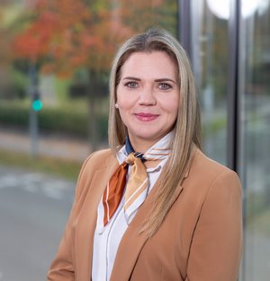 Yvonne Osterc Personalreferentin Porträt einer lächelnden Frau mit hellem langen Haar, die einen beigen Blazer und ein farbiges Halstuch trägt, vor einem Fenster mit Blick auf eine Straße und herbstliche Bäume.