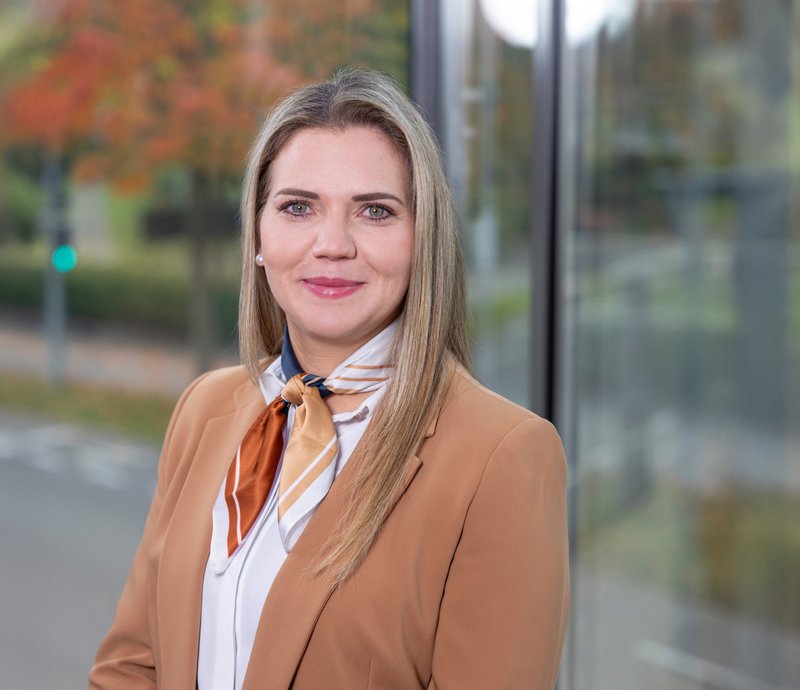 Yvonne Osterc Personalreferentin Porträt einer lächelnden Frau mit hellem langen Haar, die einen beigen Blazer und ein farbiges Halstuch trägt, vor einem Fenster mit Blick auf eine Straße und herbstliche Bäume.