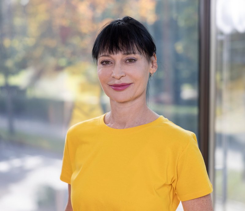 Tamara Cherednichenko Porträt einer lächelnden Frau mit dunklen Haaren und Pony, die ein gelbes T-Shirt trägt, vor einem Fenster mit unscharfem herbstlichem Hintergrund.