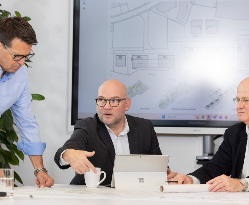 Drei Männer in einem Büro Drei Männer in einem Büro diskutieren Pläne. Im Hintergrund ist ein Bildschirm mit technischen Zeichnungen zu sehen. Einer der Männer zeigt mit der Hand auf den Tisch.