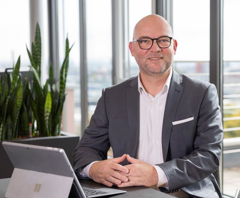 Geschäftsführer der SWG Friedrichshafen Porträt eines lächelnden Geschäftsmannes in Anzug und Brille, sitzend an einem Schreibtisch mit Laptop, vor großen Fenstern in einem modernen Büro.