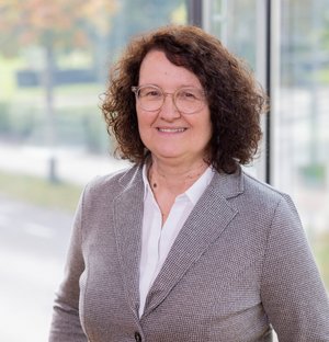 Christine Göpfert Assistenz der Geschäftsführung Porträt einer lächelnden Frau mittleren Alters mit lockigem braunem Haar und Brille, die einen grauen Blazer und ein weißes Hemd trägt. Sie steht vor einem großen Fenster mit unscharfem Hintergrund.