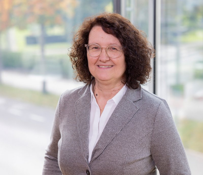 Christine Göpfert Assistenz der Geschäftsführung Porträt einer lächelnden Frau mittleren Alters mit lockigem braunem Haar und Brille, die einen grauen Blazer und ein weißes Hemd trägt. Sie steht vor einem großen Fenster mit unscharfem Hintergrund.