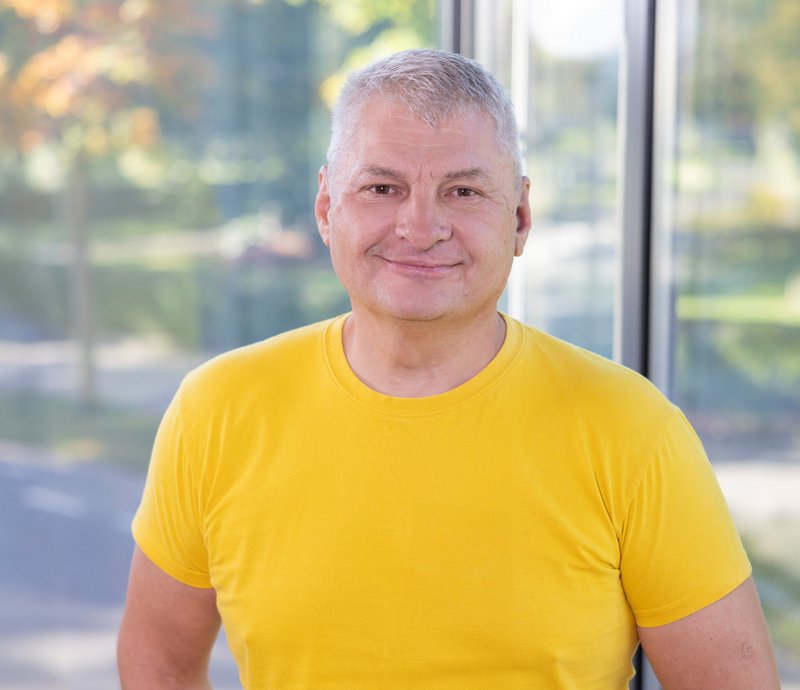 Regiebetrieb – André Hildeslev Porträt eines lächelnden älteren Mannes mit grauen Haaren, der ein gelbes T-Shirt trägt, vor einem Fenster mit verschwommenem Hintergrund.