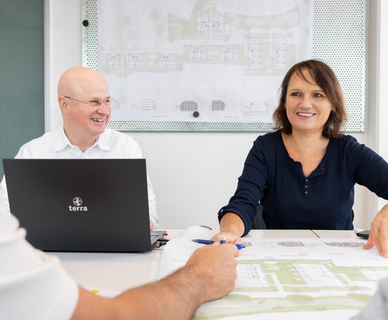 Besprechung im Team Eine Gruppe von Menschen sitzt gemeinsam an einem Tisch und betrachtet Baupläne, während sie sich austauschen.