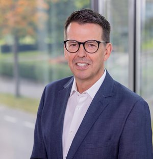 Dieter Hoffmann Leiter kaufmännische Abteilung/Prokurist Porträt eines lächelnden Mannes mittleren Alters mit dunkler Brille, weißem Hemd und blauem Anzug, stehend vor einem Fenster mit unscharfem Außenblick.
