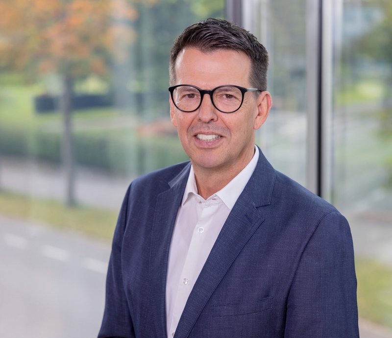 Dieter Hoffmann Leiter kaufmännische Abteilung/Prokurist Porträt eines lächelnden Mannes mittleren Alters mit dunkler Brille, weißem Hemd und blauem Anzug, stehend vor einem Fenster mit unscharfem Außenblick.