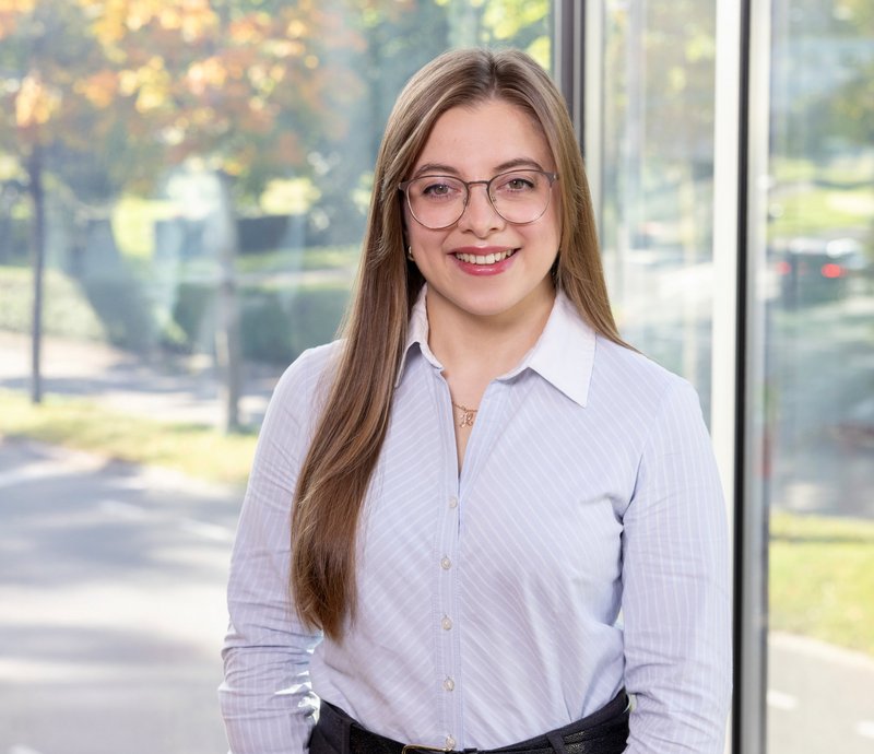 Assistenz Mieterservice – Nadlia Dossinger Junge Frau mit langer, glatter blonder Haare und Brille steht lächelnd vor einem großen Fenster mit Blick auf einen herbstlichen Park. Sie trägt ein weißes Hemd und schwarze Hose.