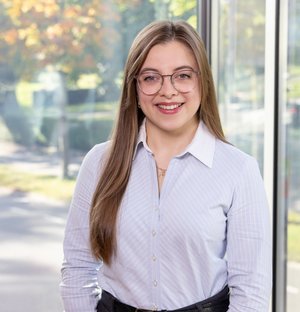 Assistenz Mieterservice – Nadlia Dossinger Junge Frau mit langer, glatter blonder Haare und Brille steht lächelnd vor einem großen Fenster mit Blick auf einen herbstlichen Park. Sie trägt ein weißes Hemd und schwarze Hose.