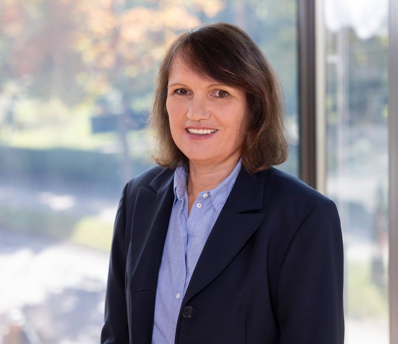 Elke Krost Assistenz Planen und Bauen Porträt einer lächelnden Frau mit braunen Haaren, die formelle Kleidung trägt, vor einem Fenster mit unscharfem Außenbereich im Hintergrund.