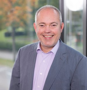 Matthias Buck Leiter Mieterservice Porträt eines lächelnden Mannes mittleren Alters, der einen hellen Hemdkragen unter einem grauen Sakko trägt.