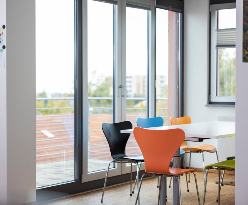 Besprechungsraum bei der SWG Ein heller, moderner Besprechungsraum mit großem Fenster, mehreren farbigen Stühlen um einen weißen Tisch und Blick auf Dächer und Bäume draußen.