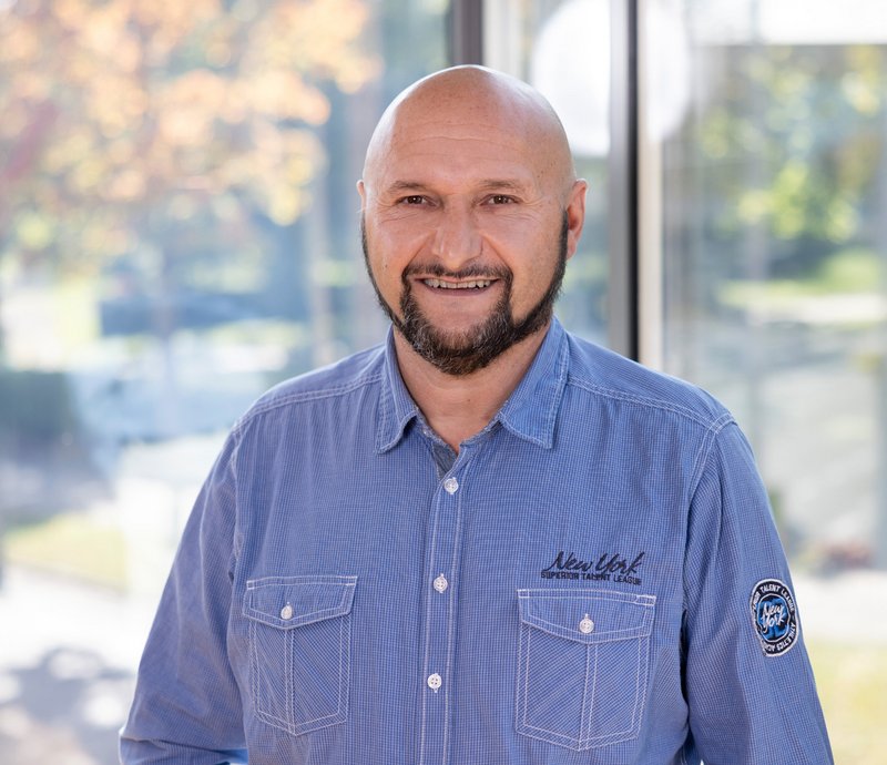 Wolfgang Ammann Haustechnik Mann mittleren Alters mit Glatze und Bart, der freundlich in die Kamera lächelt, trägt ein blaues Hemd, stehend vor einem unscharfen, hellen Hintergrund mit Bäumen.