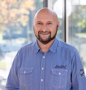 Wolfgang Ammann Haustechnik Mann mittleren Alters mit Glatze und Bart, der freundlich in die Kamera lächelt, trägt ein blaues Hemd, stehend vor einem unscharfen, hellen Hintergrund mit Bäumen.