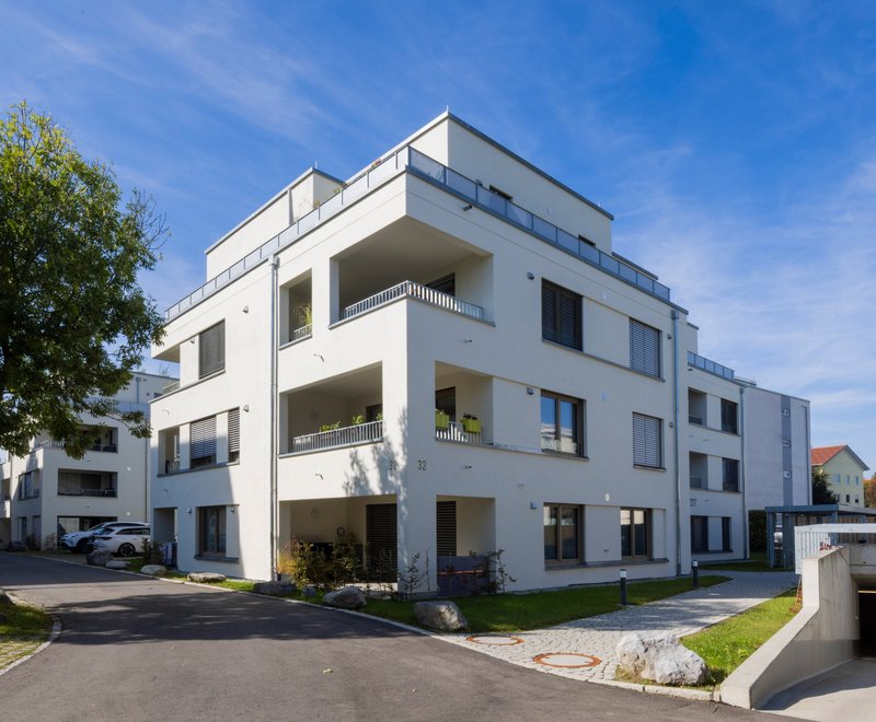 Moderner, weißer Wohnkomplex Moderner, weißer Wohnkomplex mit mehreren Stockwerken, Balkonen und umgeben von Grünflächen, unter blauem Himmel. Rechts Eingang zur Tiefgarage, links Parkplätze und Bäume.
