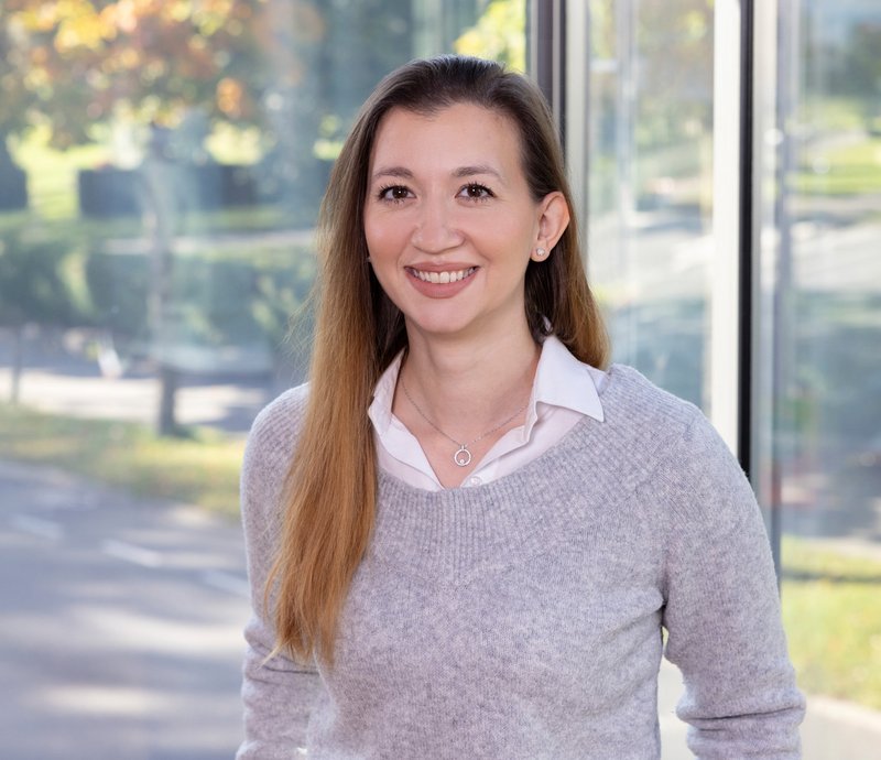 Portrait Michèle Heuser Junge Frau mit langem braunem Haar und grauem Pullover lächelt freundlich, im Hintergrund ist ein sonniger Park durch ein Fenster zu sehen.