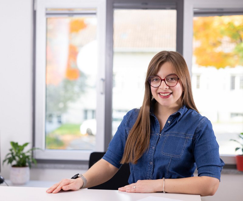 Beratung mit Herz und Kompetenz Junge Frau mit langem Haar und Brille lächelt freundlich, sitzt an einem weißen Schreibtisch vor einem großen Fenster mit Blick nach draußen, im hellen, modernen Raum.