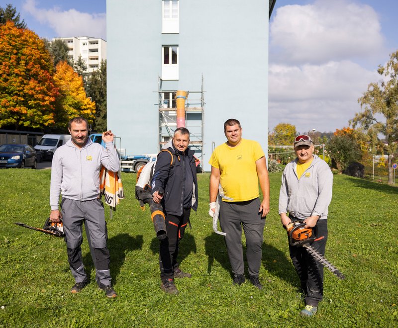Gemeinsam für Friedrichshafen Vier Männer stehen auf einer Wiese vor einem Wohngebäude, tragen Arbeitskleidung und halten Gartengeräte, bereit für Garten- oder Landschaftspflegearbeiten.