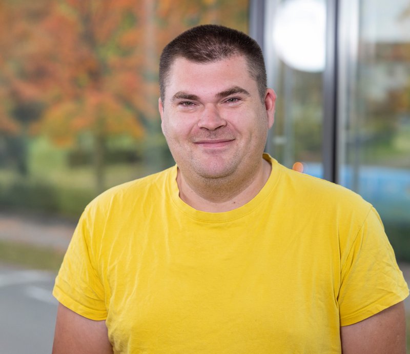 Mykola Hrom Ein Mann mit kurzem braunem Haar und gelbem T-Shirt lächelt in die Kamera. Im Hintergrund sind unscharfe Herbstbäume und ein Fenster zu sehen.