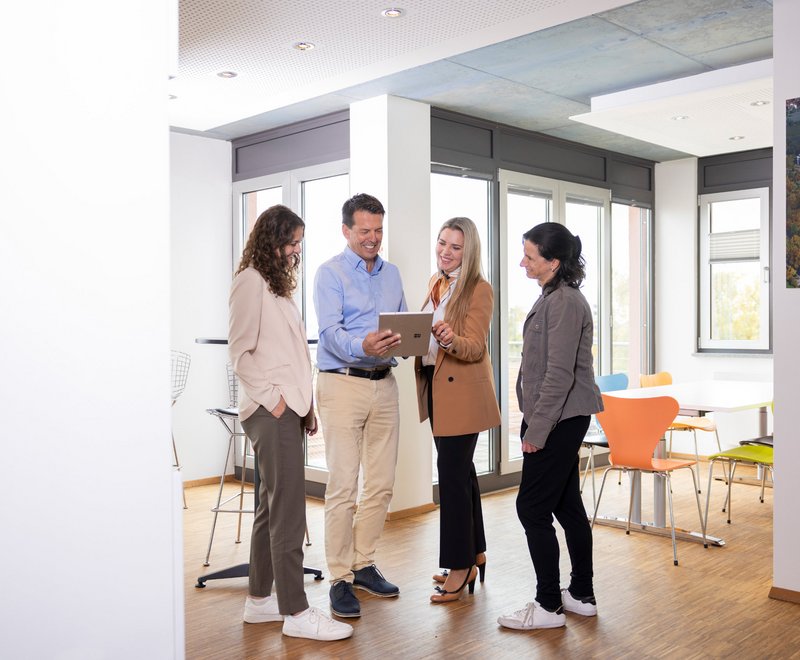 Besprechung in der Räumlichkeit der SWG Vier Personen stehen in einem modernen Büro in einer Gruppe zusammen. Ein Mann zeigt den anderen drei mit einem Tablet etwas und alle wirken interessiert und freundlich.