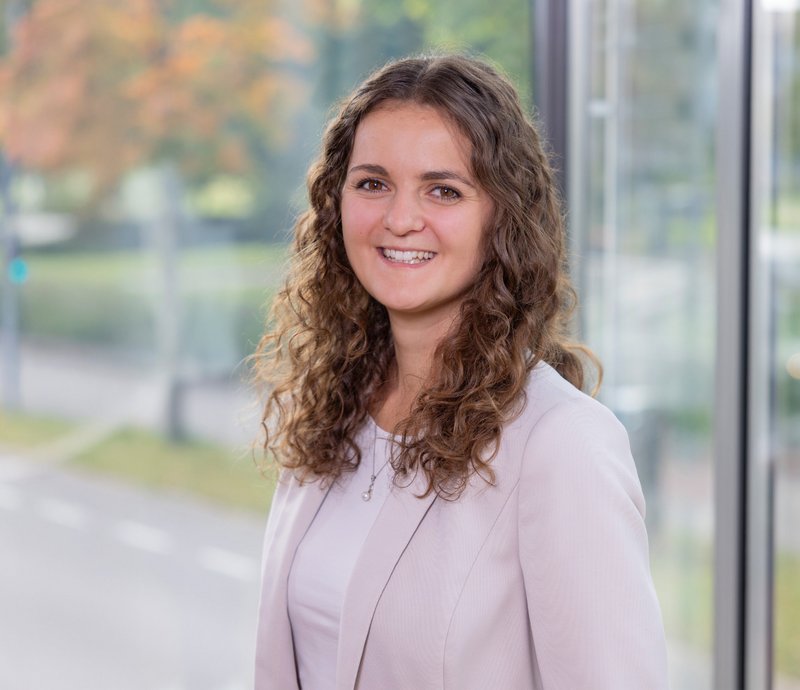 Jessica Spieler Projektmanagement Stabsstelle Junge Frau mit lockigem Haar und hellem Blazer lächelt freundlich in die Kamera, vor unscharfem herbstlichem Hintergrund und moderner Glasfassade.