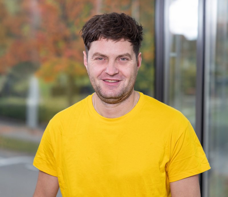 Hryhoii Babenko Porträt eines lächelnden Mannes mit kurzen dunklen Haaren, der ein gelbes T-Shirt trägt. Unscharfer Hintergrund mit Herbstbäumen und Gebäudefassade.