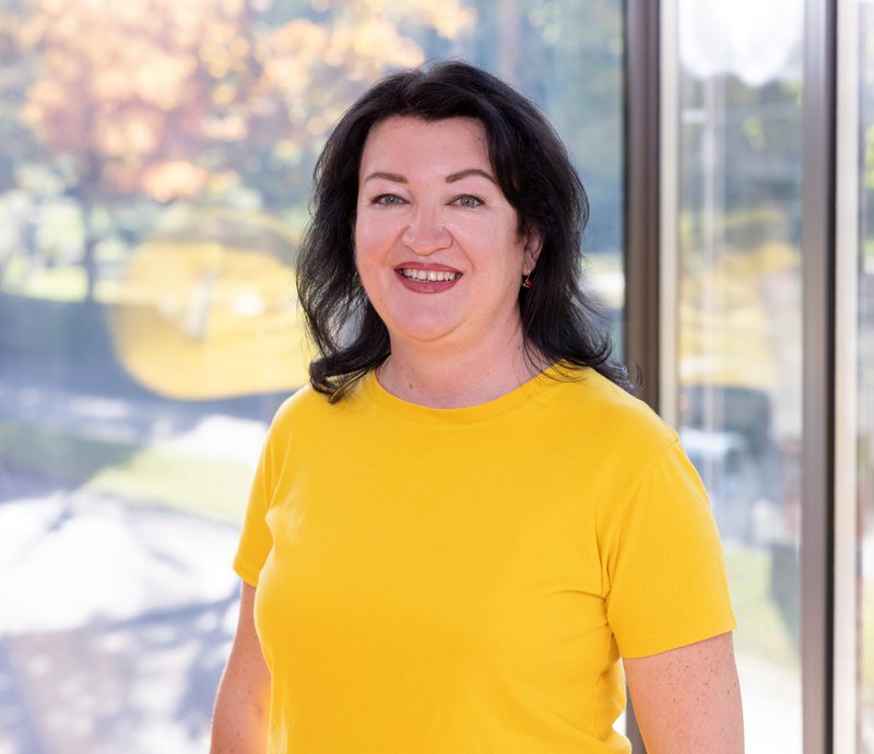 Tetiana Otrishchenko Portrait einer lächelnden Frau mit dunklem Haar, die ein gelbes T-Shirt trägt, vor einem Fenster mit Blick auf eine herbstliche Landschaft.