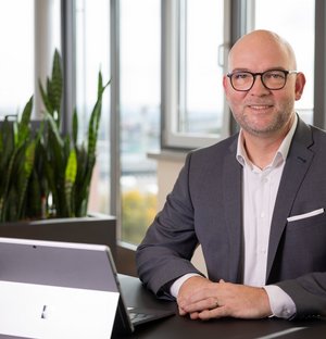 Geschäftsleitung der SWG Friedrichshafen Ein lächelnder Mann mit Brille und weißem Hemd steht neben einem Fenster, die Arme locker an den Seiten. Die Umgebung wirkt hell und freundlich.