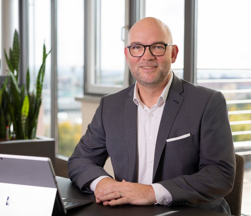 Geschäftsleitung der SWG Friedrichshafen Ein lächelnder Mann mit Brille und weißem Hemd steht neben einem Fenster, die Arme locker an den Seiten. Die Umgebung wirkt hell und freundlich.