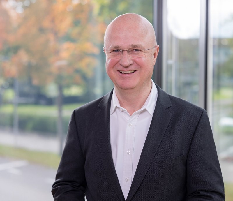 Rudolf Hilebrand SWG Friedrichshafen Porträt eines lächelnden, älteren Mannes mit Brille und Glatze, der einen dunklen Anzug und ein weißes Hemd trägt.