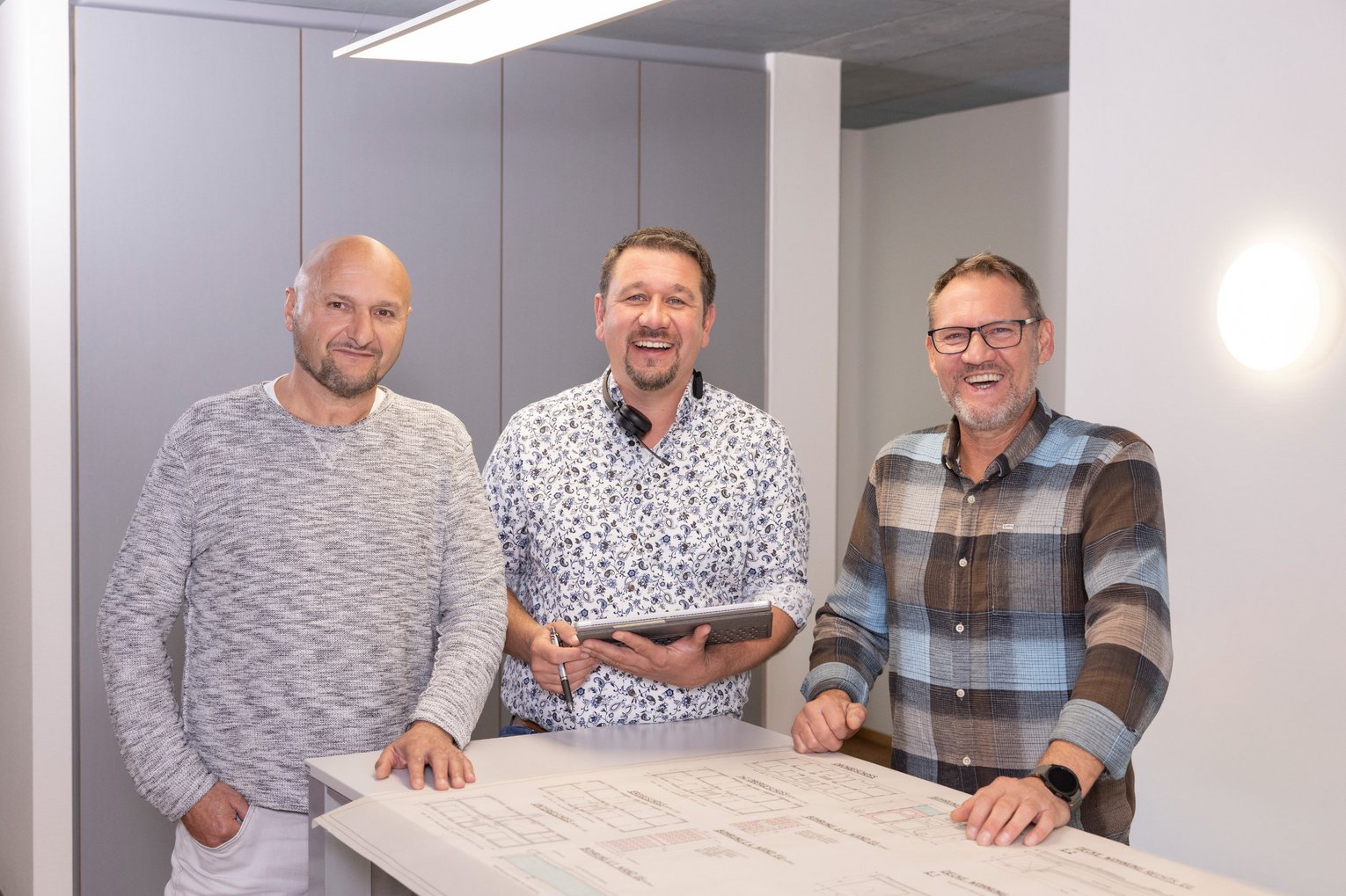 SWG Team Schadensmeldung Drei lächelnde Männer stehen gemeinsam an einem großen Tisch mit Unterlagen und wirken wie in einer Besprechung oder Zusammenarbeit.