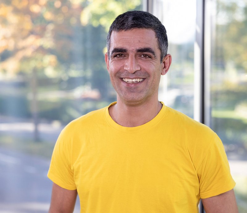 Regiebetrieb – Mohammad Salimi Porträt eines lächelnden Mannes mit kurzen dunklen Haaren und gelbem T-Shirt vor unscharfem, hellem Hintergrund im Freien.