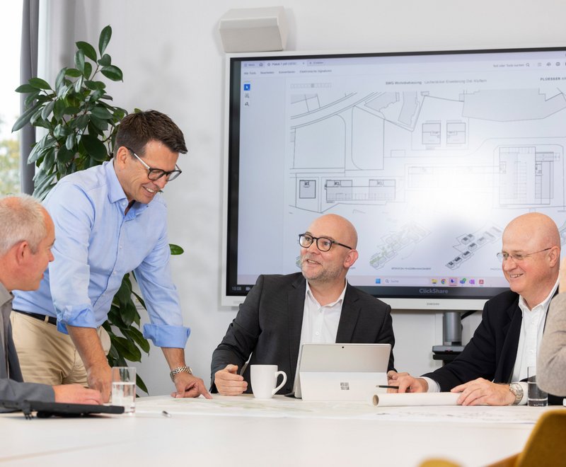 Besprechung bei der SWG Eine Gruppe von fünf Personen sitzt in einem Besprechungsraum um einen Tisch und diskutiert vor einem großen Bildschirm mit technischen Zeichnungen oder Plänen. Alle wirken konzentriert und freundlich im Austausch.