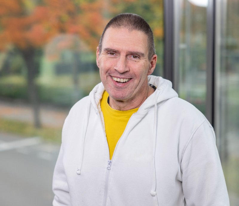 Harald Reiner Ein lächelnder, mittelalter Mann in einem weißen Kapuzenpullover und gelbem T-Shirt steht vor einem herbstlichen Hintergrund mit Bäumen und Straße.