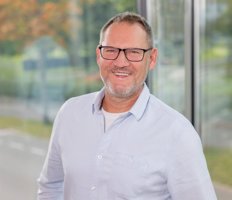 Armin Wahr Technischer Mieterservice Porträt eines lächelnden mittelalten Mannes mit Brille, der ein hellblaues Hemd trägt. Er steht vor einem großen Fenster mit unscharfem Hintergrund.
