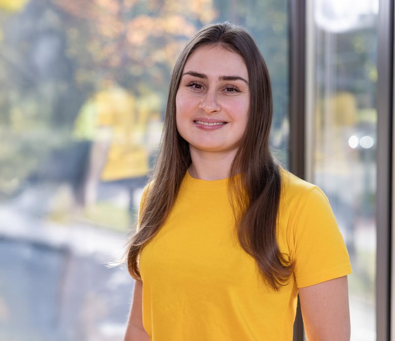 Hanna Hrom Junge Frau mit langen braunen Haaren und gelbem T-Shirt steht lächelnd vor einem Fenster.