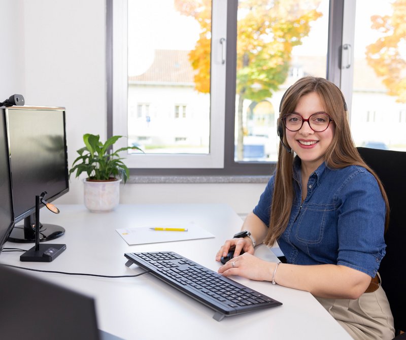 Arbeitsplatz mit Ausblick Junge Frau sitzt lächelnd am Schreibtisch vor einem Computer mit zwei Bildschirmen in einem hellen Büro.