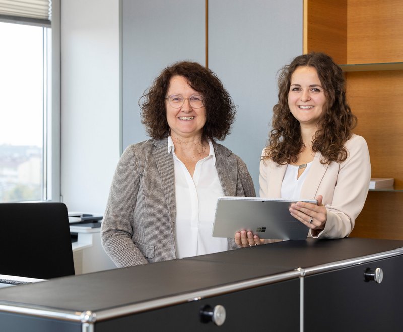 Büromitarbeiterinnen SWG Zwei lächelnde Frauen stehen in einem modernen Büro hinter einem Schreibtisch, eine hält ein Tablet, die andere neben ihr.