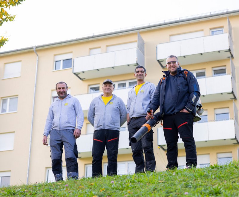 SWG-Team vor dem Bürogebäude Vier Männer in Arbeitskleidung stehen vor einem Wohnhaus und lächeln. Einer hält ein Laubbläsergerät. Sie wirken bereit für Garten- oder Hauswartungsarbeiten.
