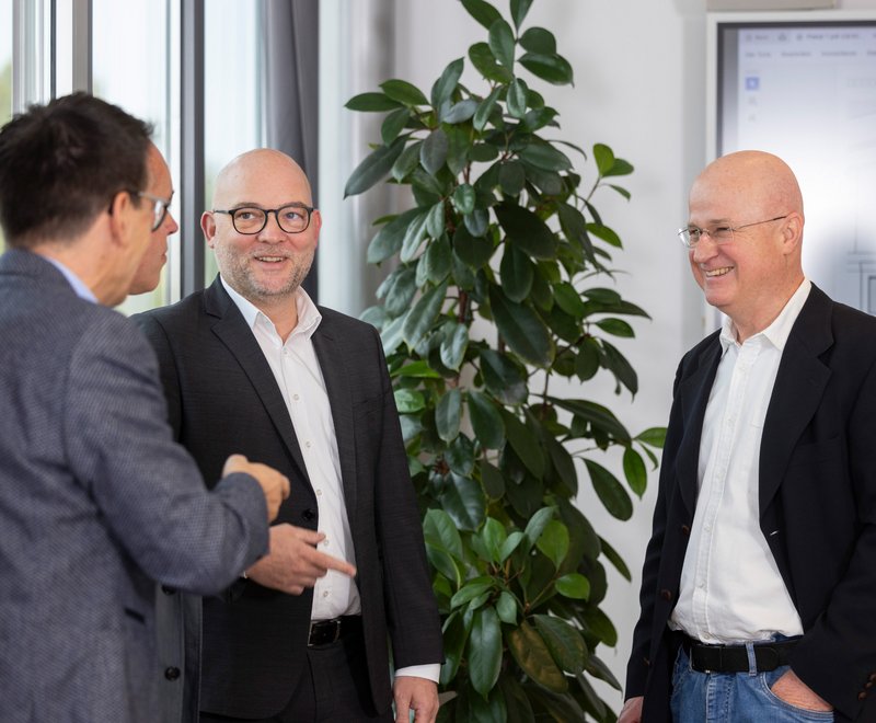 Mitarbeiter der SWG im Gespräch Drei Männer im Anzug stehen in einem Büro und unterhalten sich freundlich miteinander. Zwei von ihnen lächeln und einer gestikuliert während des Gesprächs. Im Hintergrund steht eine große grüne Pflanze.