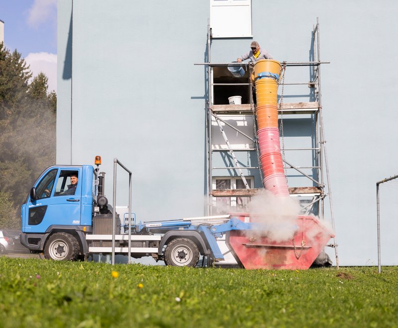 Wohnquartier der SWG am Bodensee Ein Bauarbeiter steht auf einem Gerüst an einem Wohnhaus und wirft Bauschutt mit einer langen, bunten Rutsche in einen großen Container auf einem kleinen Lastwagen, der vor dem Gebäude steht. Staub steigt während der Entsorgung auf.